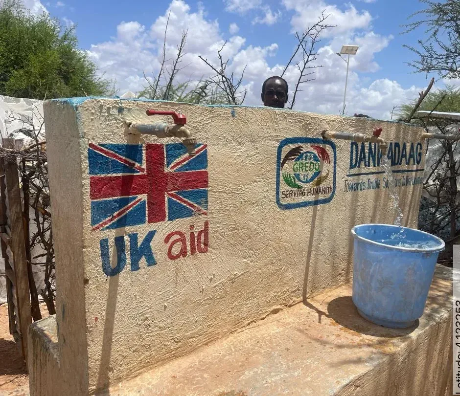 A masonry tap stand painted with UK aid, GREDO and Danwadaag markings, its taps running into household buckets.