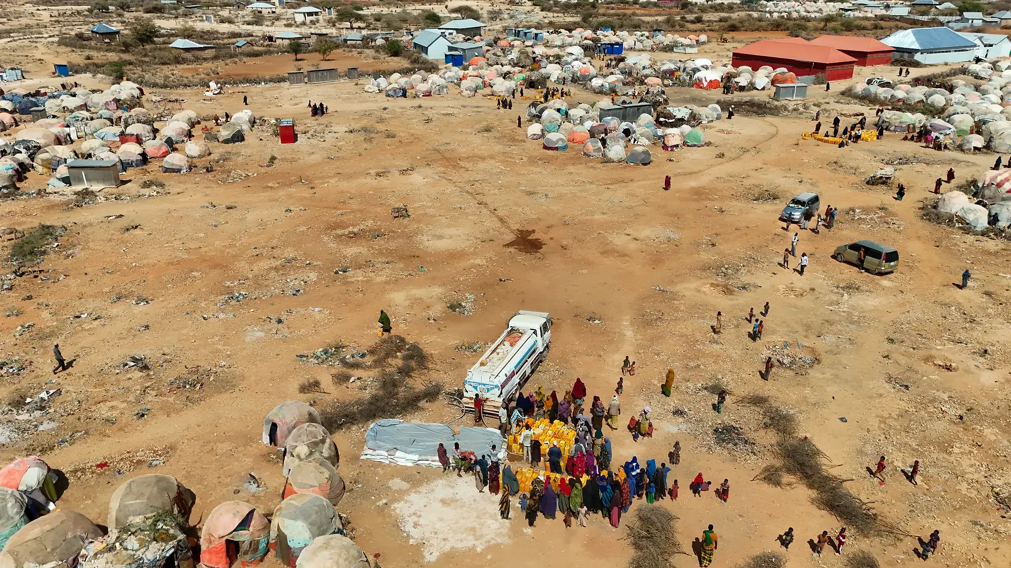 Aerial view of a settlement edge, dome shelters and fenced family plots on bare earth with tin-roofed buildings and a red-roofed compound in the distance.