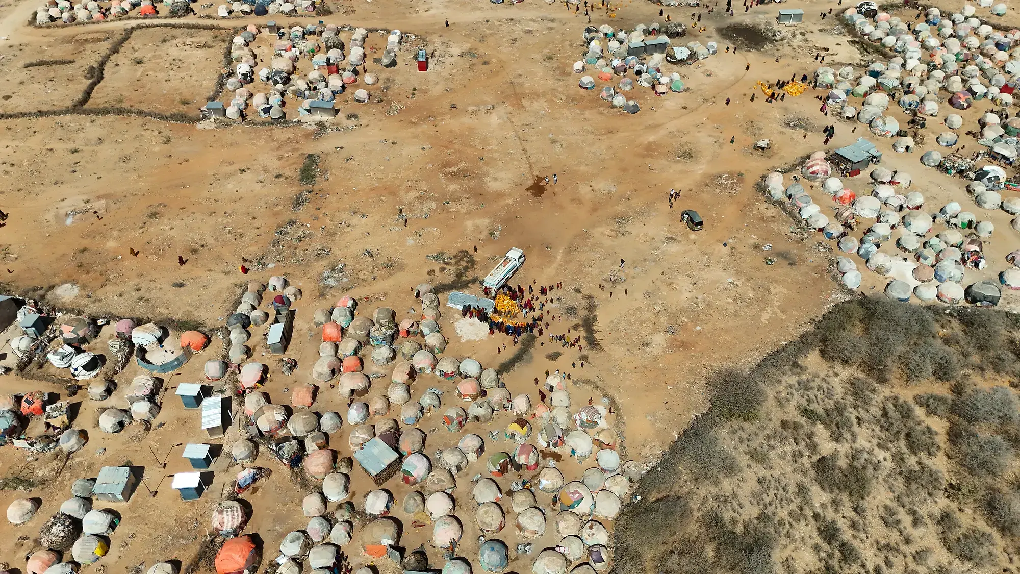High aerial view over a displacement settlement, clusters of dome shelters and walled plots around open ground, with a water tanker distribution small at its centre.