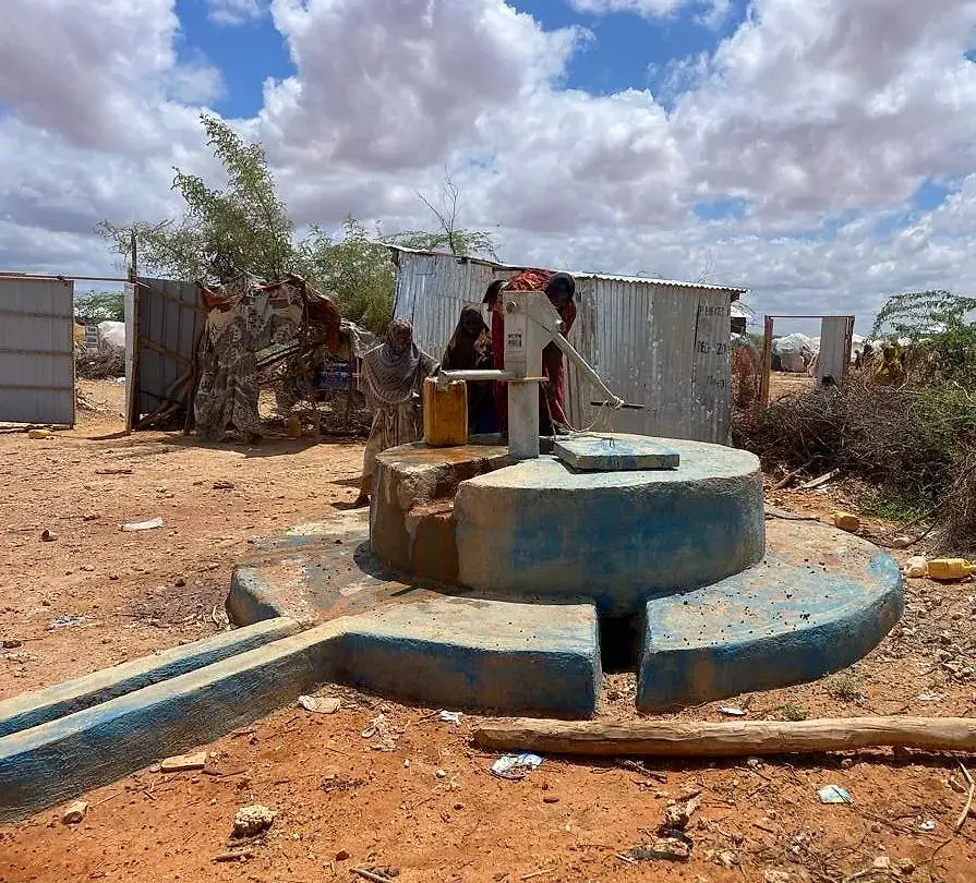 A painted masonry well head with a hand pump, women collecting water into jerrycans, and corrugated-iron shelters and tents behind.