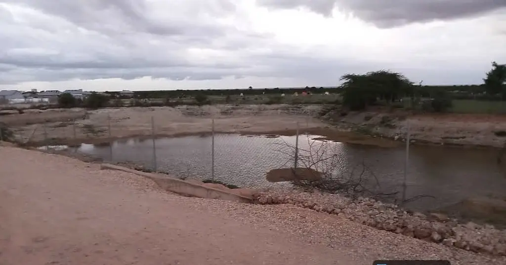A large earth catchment holding brown water behind a fence line, under a heavy grey sky.