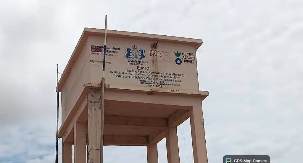 A donor-marked elevated water tank on concrete legs inside a fenced borehole compound, with a generator building beside it.