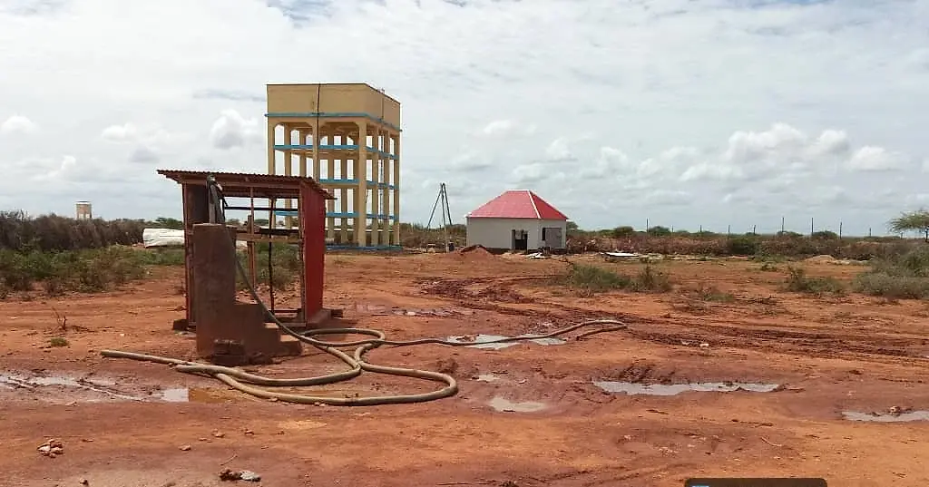 A steel pump frame and hoses in the foreground on wet red earth, with an unfinished multi-storey water tower and a red-roofed building behind.