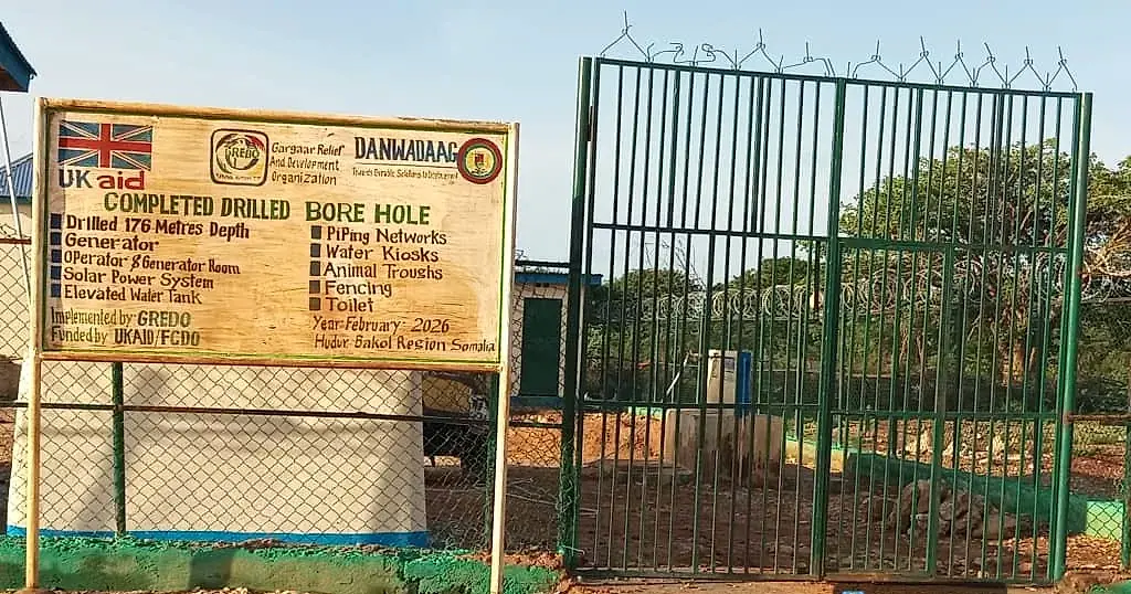A painted signboard reading Completed Drilled Bore Hole, listing the components built, in front of a fenced compound with a green steel gate.