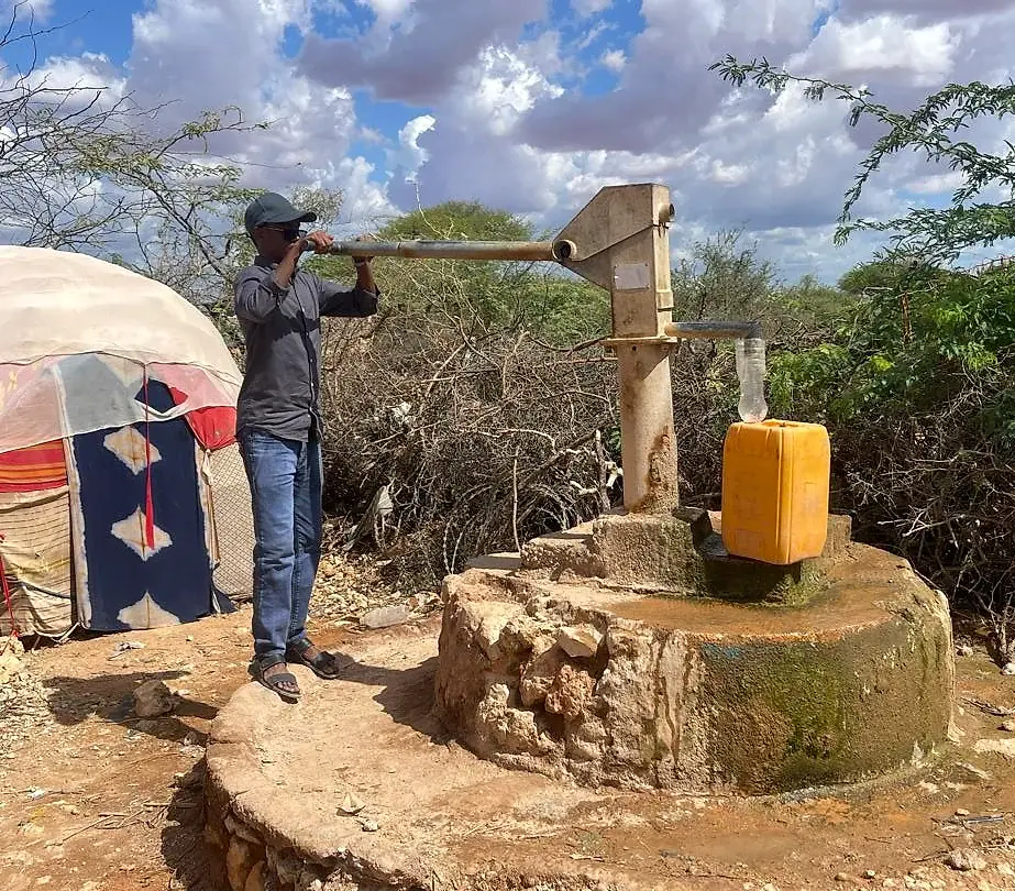 A man working the handle of a hand pump mounted on a raised masonry apron, with a yellow jerrycan filling beside him and a shelter behind.
