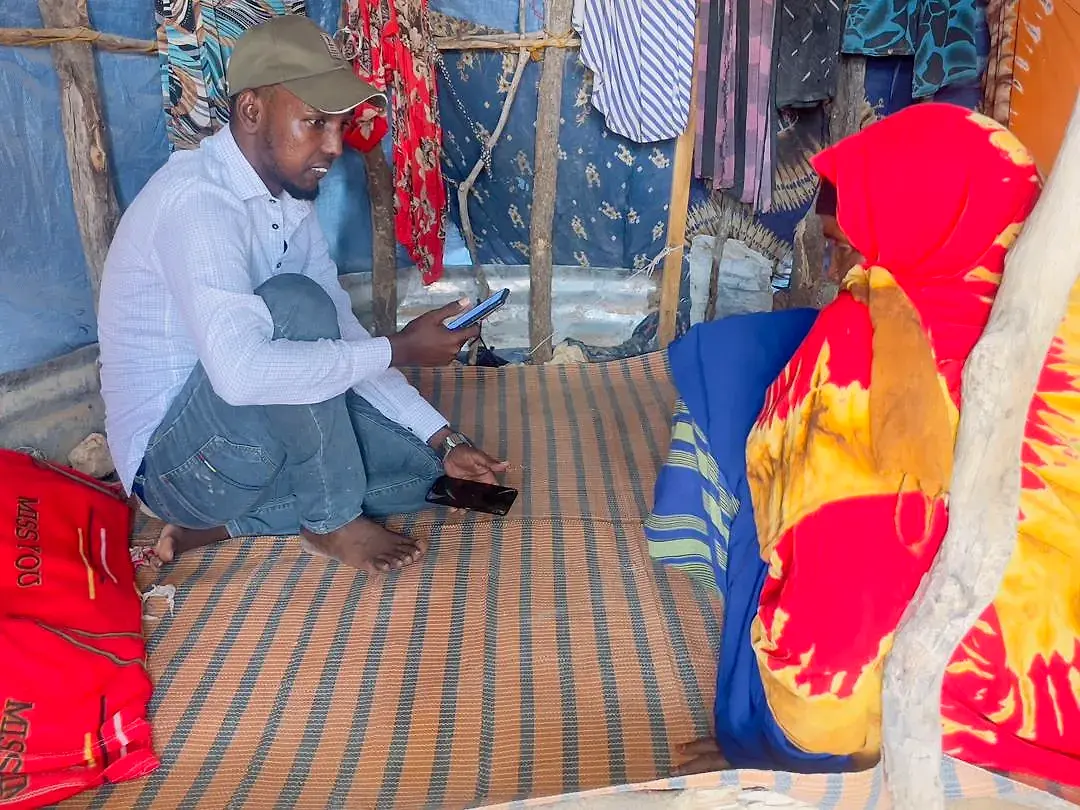 An enumerator seated on a mat inside a shelter, phones in hand, speaking with a woman seated across from him.