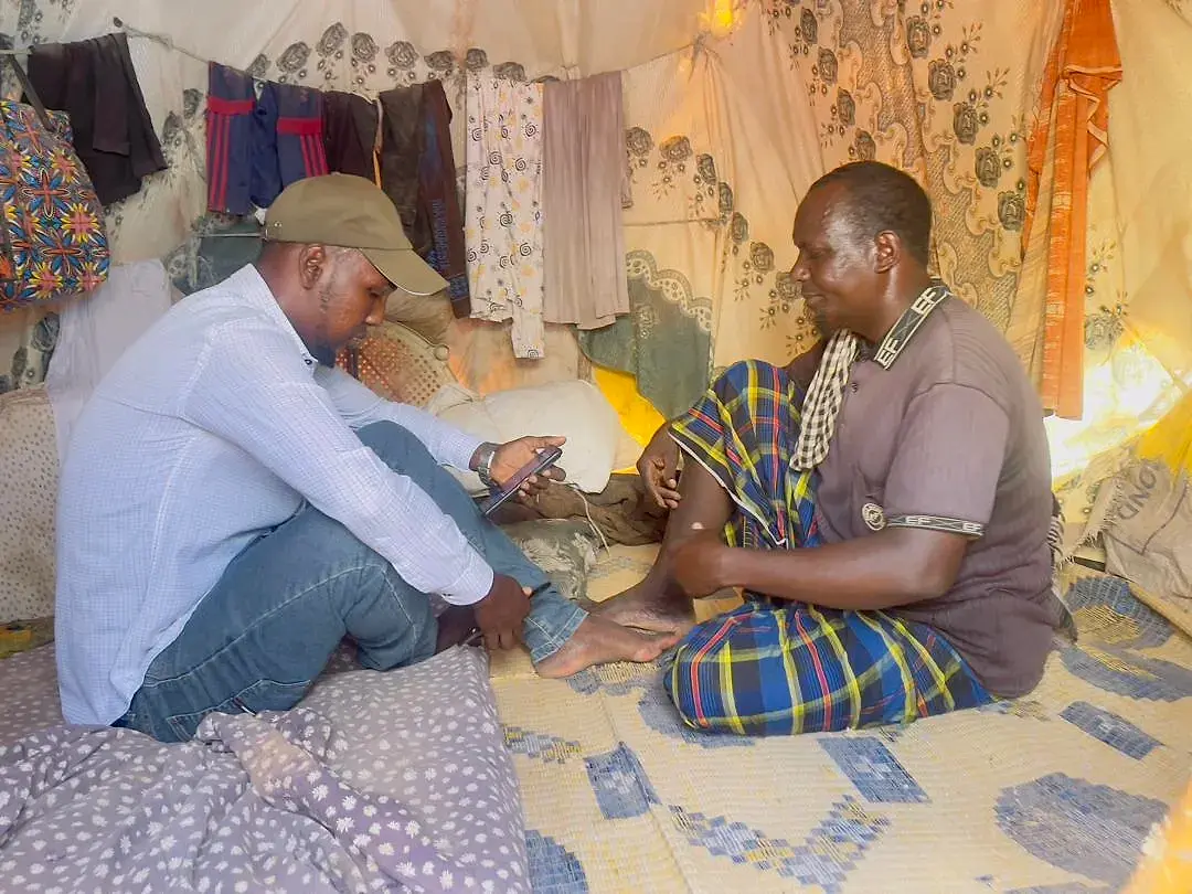 An enumerator seated on the floor of a shelter recording answers on a phone, interviewing a man seated across from him with yellow jerrycans behind.
