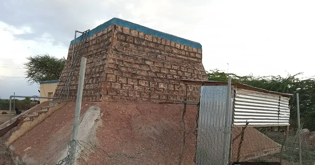 A stone-built structure part way through construction, with a corrugated iron sheet leaning against one wall and excavated earth in front.