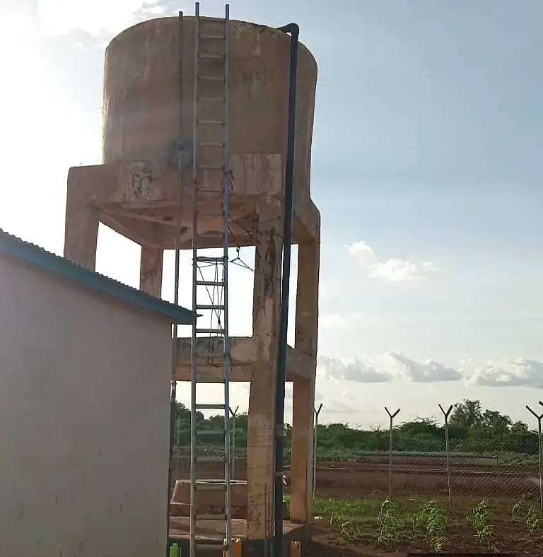 An elevated concrete storage tank with a steel ladder, beside a green-roofed building inside a fenced compound.