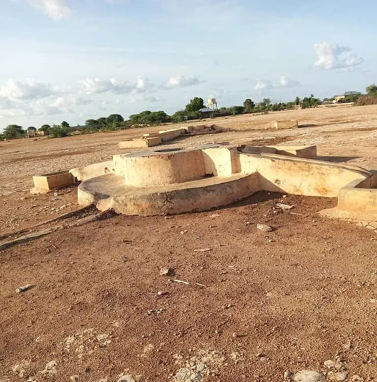 A cluster of masonry shallow well heads and troughs on open ground, with trees and an elevated tank on the horizon.