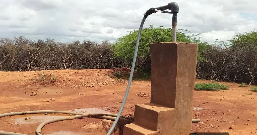 A masonry standpipe with a tap and a long black hose coiled across bare red earth, with thorn scrub behind.