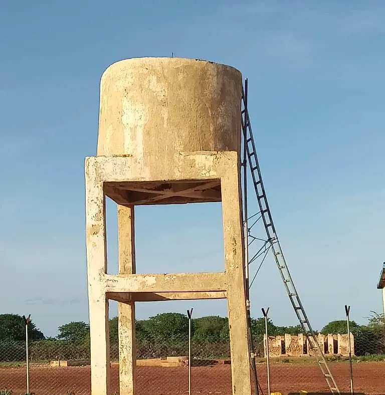 A concrete storage tank raised on four legs against a blue sky, with an access ladder and a fence line below.