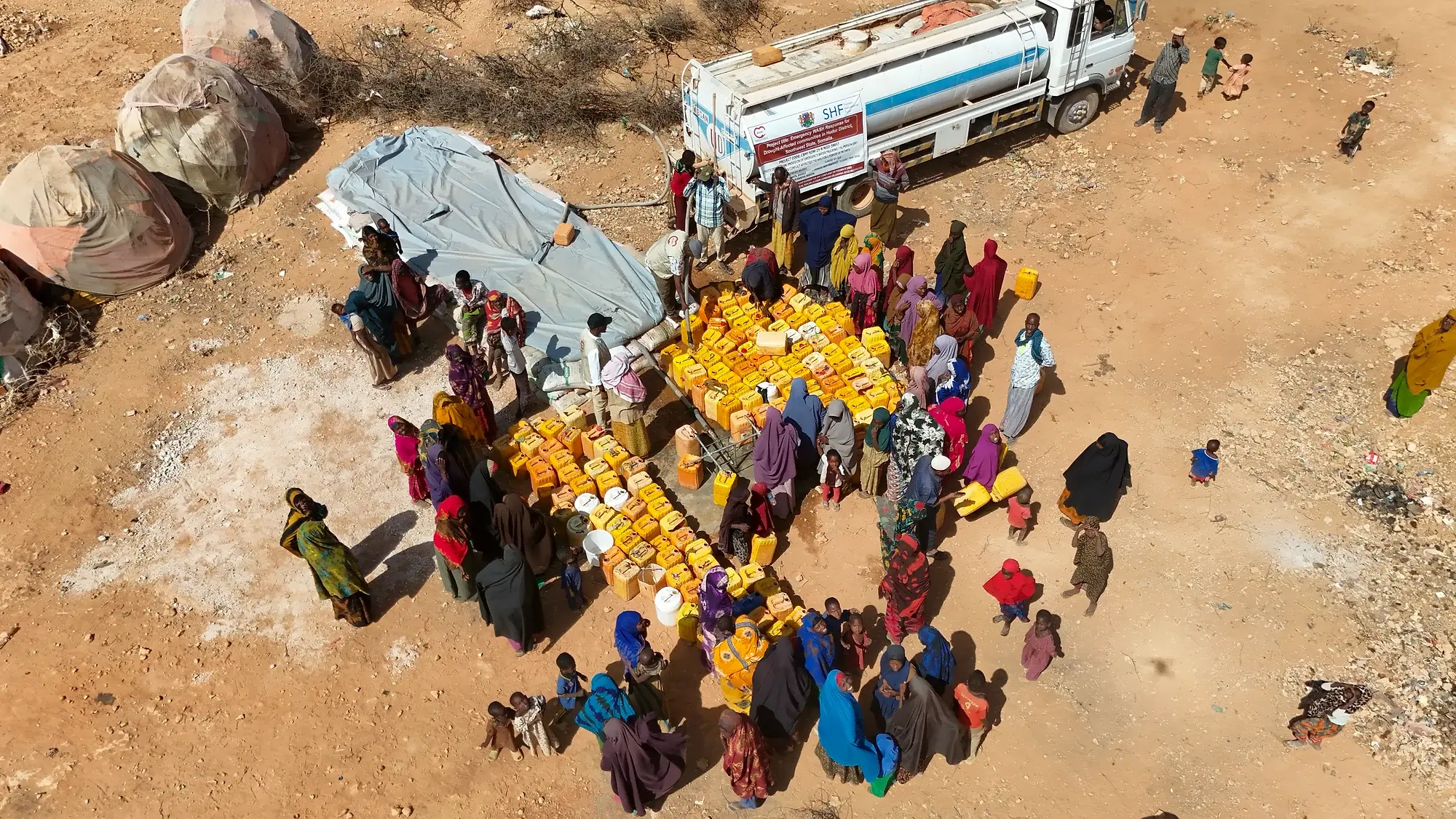 Looking down on a water tanker parked on bare ground, with a crowd of people gathered around several hundred yellow jerrycans laid out in rows on the sand.