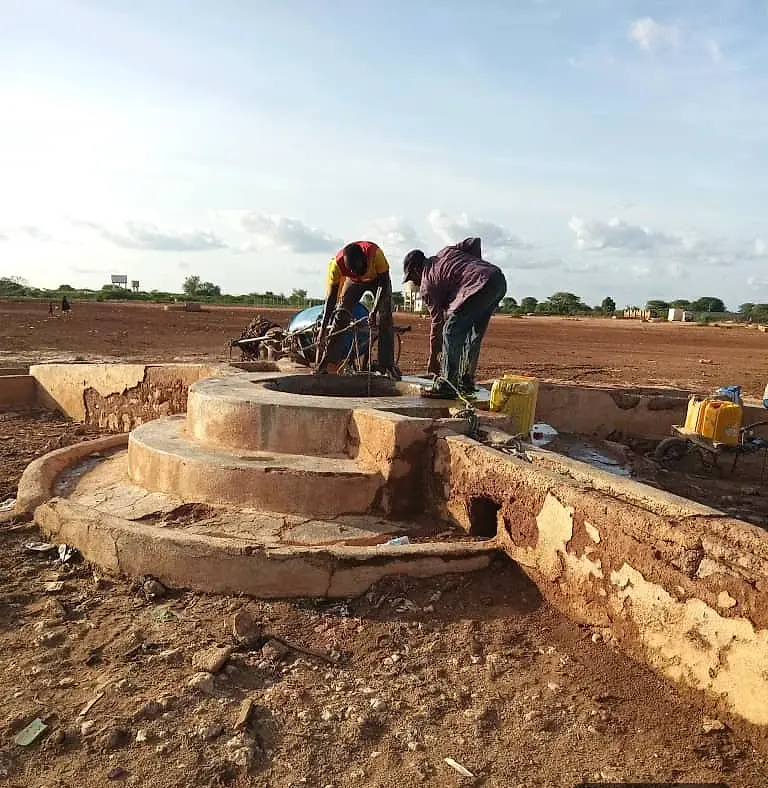 Two people working at the top of a circular concrete well head, on an apron of turned red earth with jerrycans beside them.