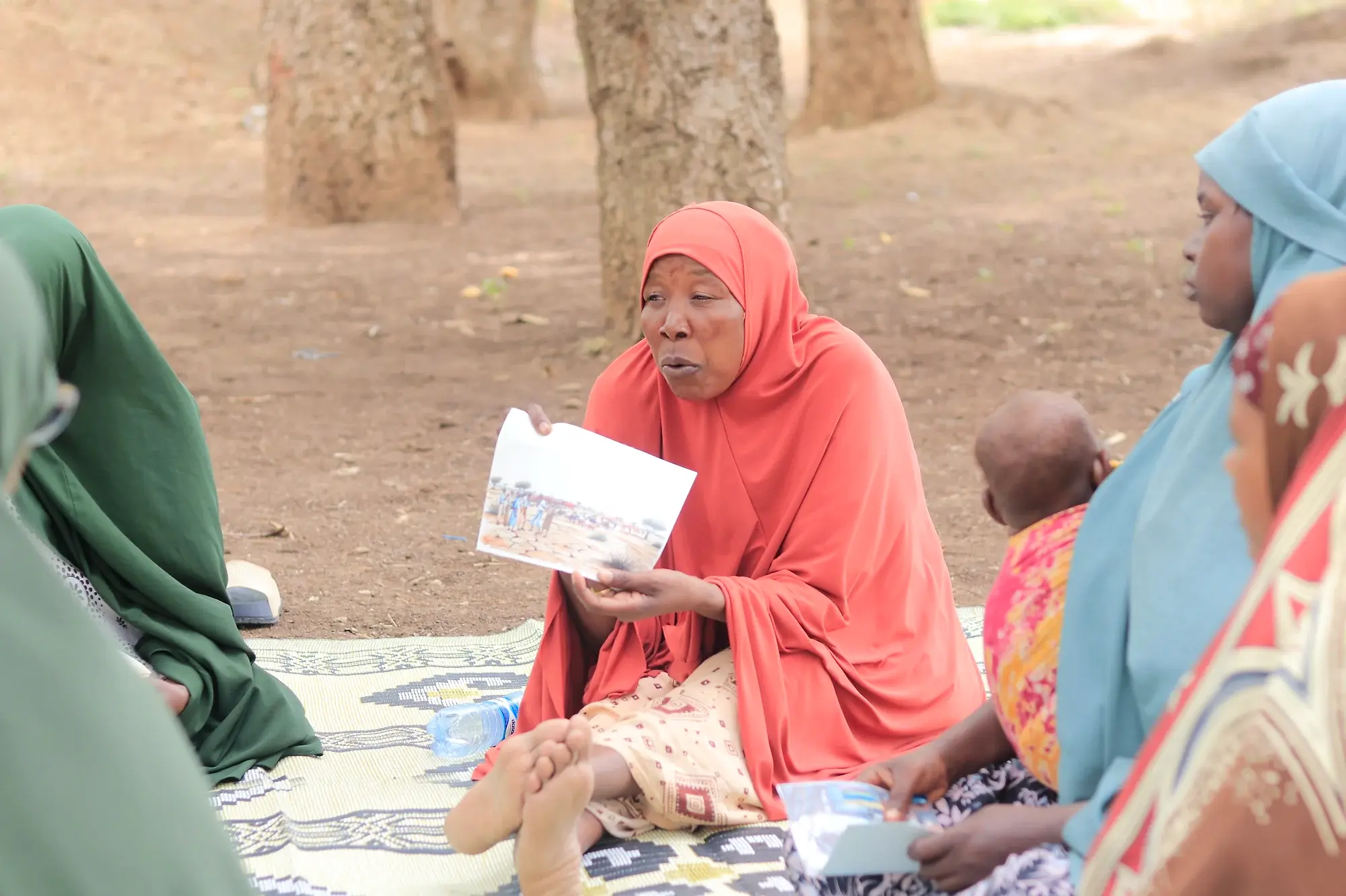 A woman speaking while holding up a printed illustration card, with other participants seated on mats around her under trees.